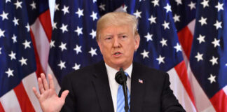 A man giving a speech with American flags behind.