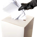 Gloved hand placing multiple papers in a ballot box.