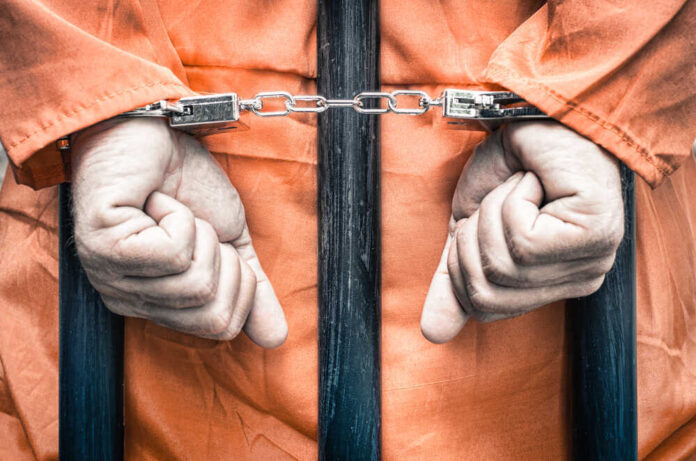 shutterstock_217094626.jpg featured image Handcuffed person in orange jumpsuit behind prison bars.
