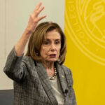 Nancy Pelosi gestures while speaking in front of yellow backdrop
