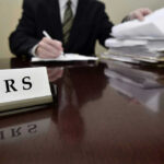IRS Shocker — Free Filing Option Axed IRS sign on desk, person handling paperwork behind.