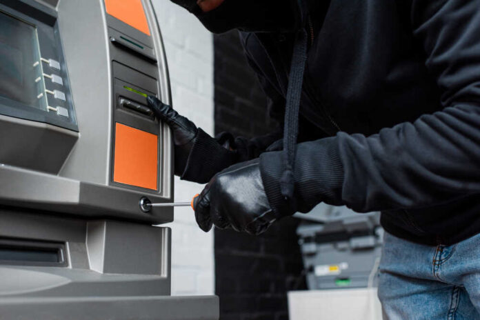 A person in a black hoodie and gloves tampering with an ATM