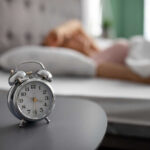 An alarm clock on a bedside table with a person sleeping in the background