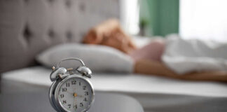 An alarm clock on a bedside table with a person sleeping in the background