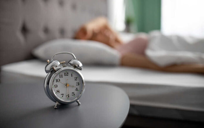 An alarm clock on a bedside table with a person sleeping in the background
