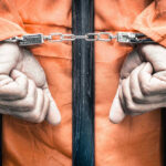 Handcuffed person in orange jumpsuit behind prison bars.