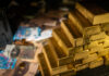 Stack of gold bars next to scattered banknotes and coins