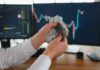 Person holding cash in front of stock market charts on computer screens