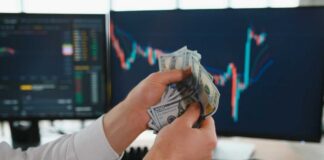 Person holding cash in front of stock market charts on computer screens