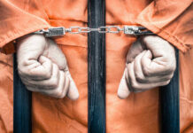 Handcuffed person in orange jumpsuit behind prison bars.