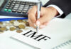 A hand writing 'IMF' on a notepad with coins and a calculator in the background