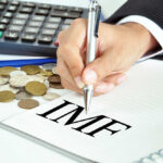 A hand writing 'IMF' on a notepad with coins and a calculator in the background