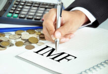 A hand writing 'IMF' on a notepad with coins and a calculator in the background