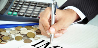 A hand writing 'IMF' on a notepad with coins and a calculator in the background