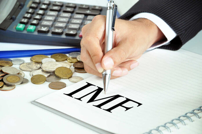 A hand writing 'IMF' on a notepad with coins and a calculator in the background