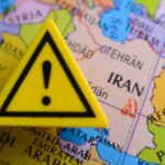 NOW: Iran Declares US Universities ‘Legitimate Targets’ A yellow warning sign placed on a map highlighting Iran