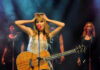 Taylor Swift holding guitar on stage, background singers behind.