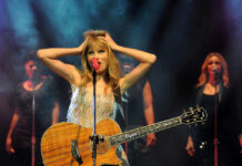 Taylor Swift holding guitar on stage, background singers behind.