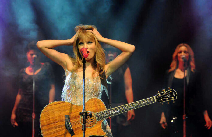 Taylor Swift holding guitar on stage, background singers behind.