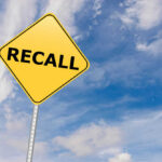 Dangerous Glitch Triggers Robotaxi Recall Yellow sign with RECALL text against blue sky.
