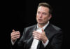 Elon Musk Dangles $1T Elon Musk speaking and gesturing with hands in discussion.