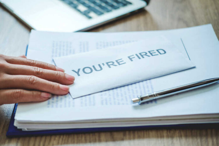 A hand resting on an envelope labeled 'YOU'RE FIRED' on a desk with papers and a pen