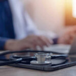 Hero Pilot’s Diagnosis Sparks Outpouring Of Support Stethoscope and tablet on desk, doctor using laptop.