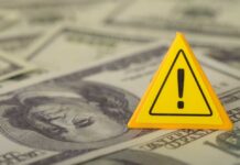 Oil Shock Slams American Wallets – AGAIN! A yellow warning sign placed on a background of dollar bills