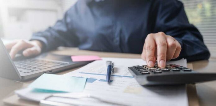 Close-up,Of,Accountant,Using,Calculator,And,Laptop,With,Financial,Documents, Person using a calculator while working on a laptop and reviewing documents