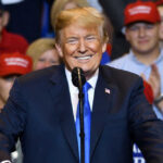 Donald Trump speaking at a rally, supporters in background.