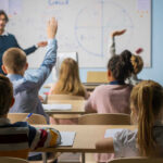 Measles Risk Spikes: Schools On Thin Ice Students raising hands in a classroom with a teacher.