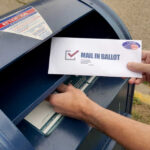 Hands mailing a ballot envelope into a blue mailbox