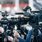 DOJ U-Turn Stuns Lincoln Pool Case Broadcast cameras on tripods at a press event
