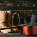 Cowboy boots, hat, and guitar on wooden surface.