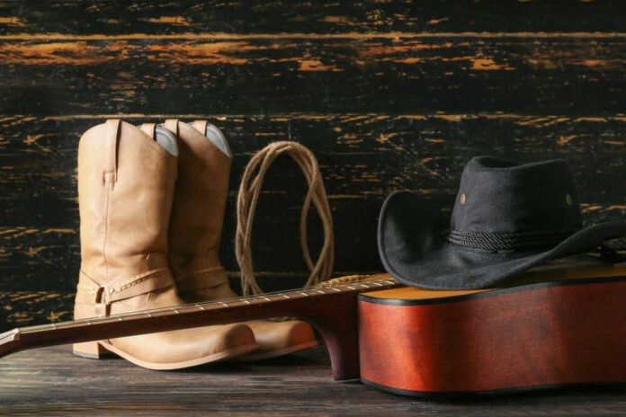 shutterstock_2338150313 Cowboy boots, hat, and guitar on wooden surface.