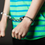 11-Year-Old Booked In Brother’s Killing Person in striped shirt wearing handcuffs.
