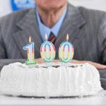 Live To 100? Drink This Trio Birthday cake with 100 candles, person in background.