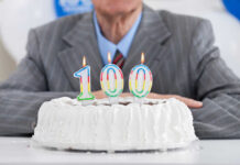 Live To 100? Drink This Trio Birthday cake with 100 candles, person in background.