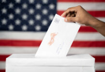 Ballots Trigger DOJ Meltdown Hand inserting mail-in ballot envelope into ballot box
