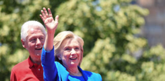 Hillary Clinton waving at an outdoor event with Bill Clinton in the background